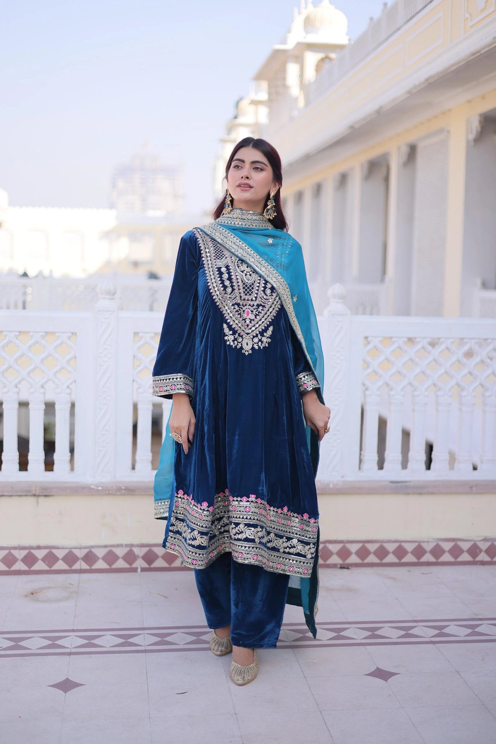 Woman in a blue traditional outfit standing on a balcony with white railings and a building in the background.