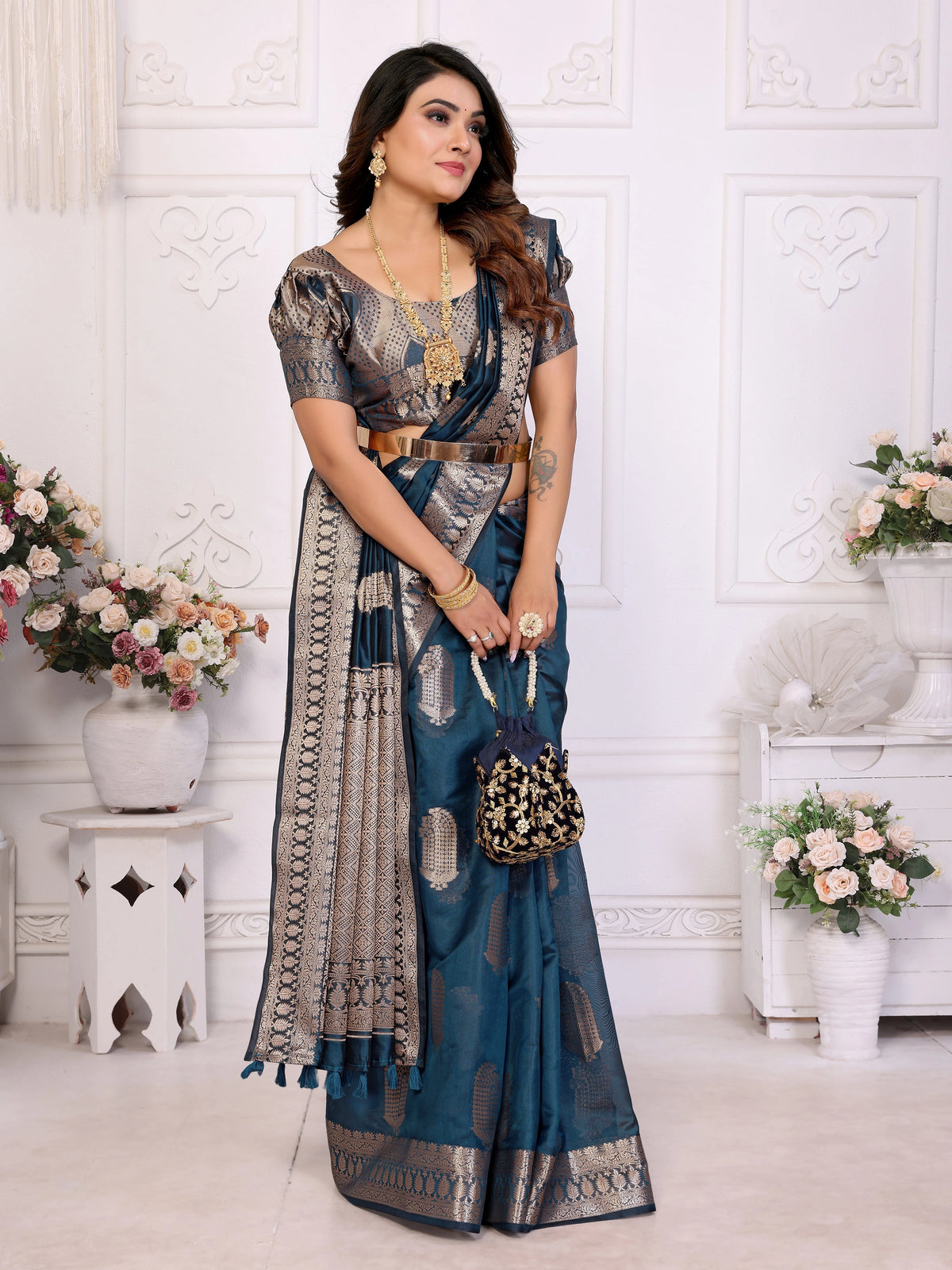 Woman in a blue and gray saree standing in a decorated room with flowers and vases.