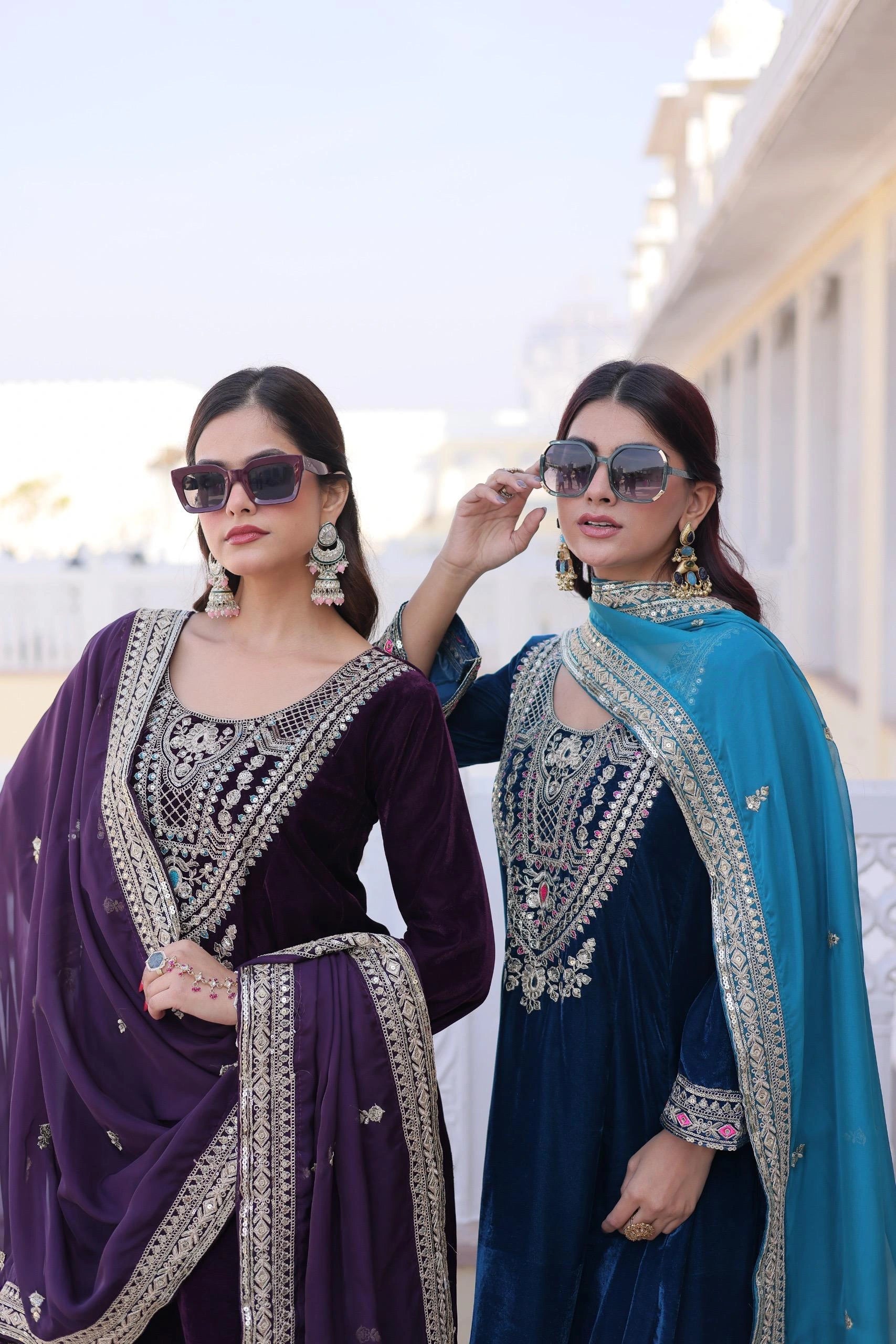 Two women wearing traditional embroidered sarees outdoors with a clear sky.