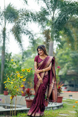 Woman in a deep purple saree with zari work standing in a garden with yellow flowers and lush greenery in the background