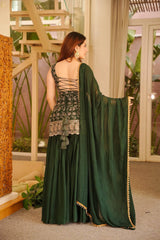 Woman in a green and silver traditional outfit standing in an indoor setting with plants and furniture.