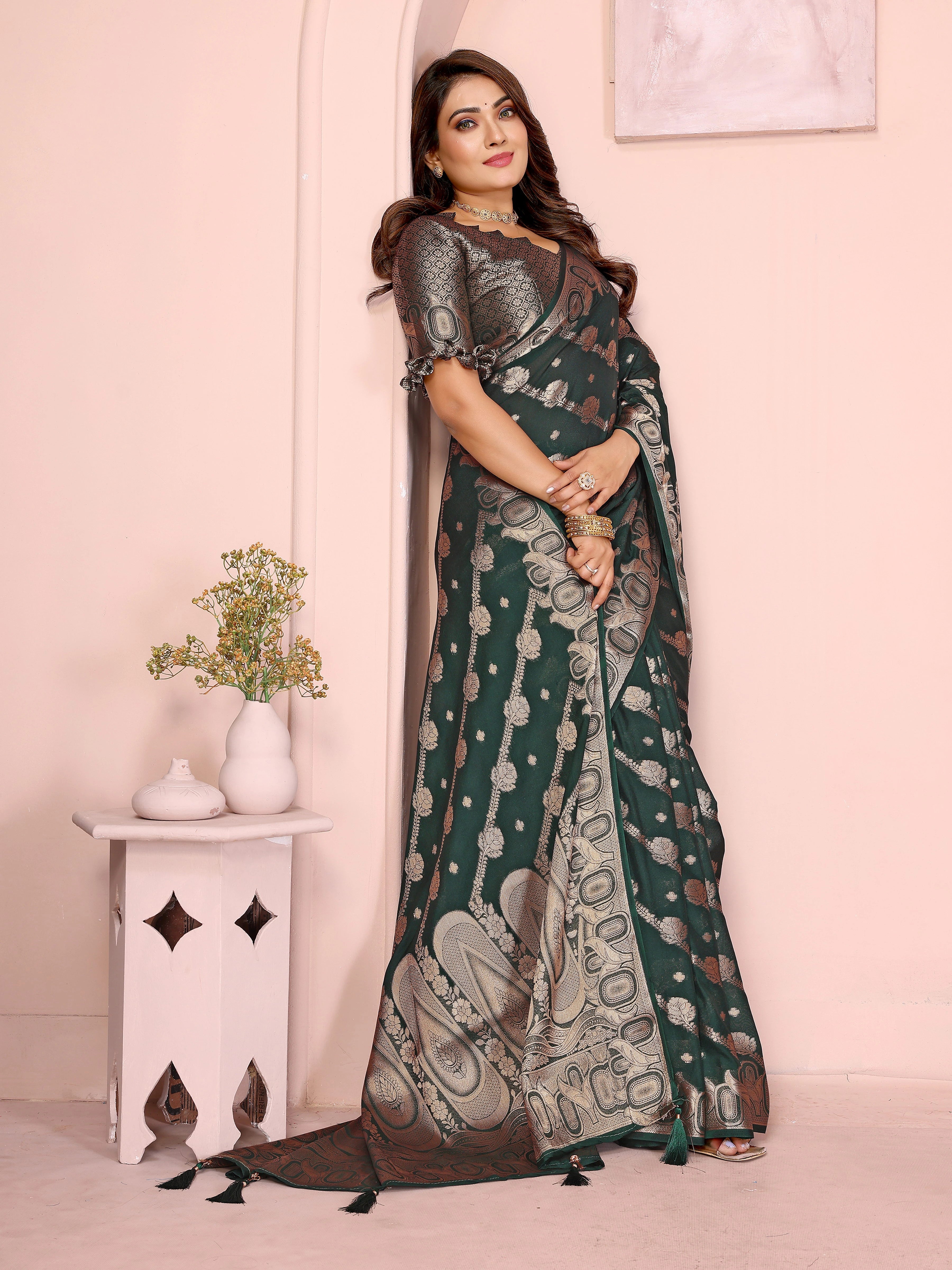 Woman wearing a dark green saree with a pattern, standing against a light pink wall.