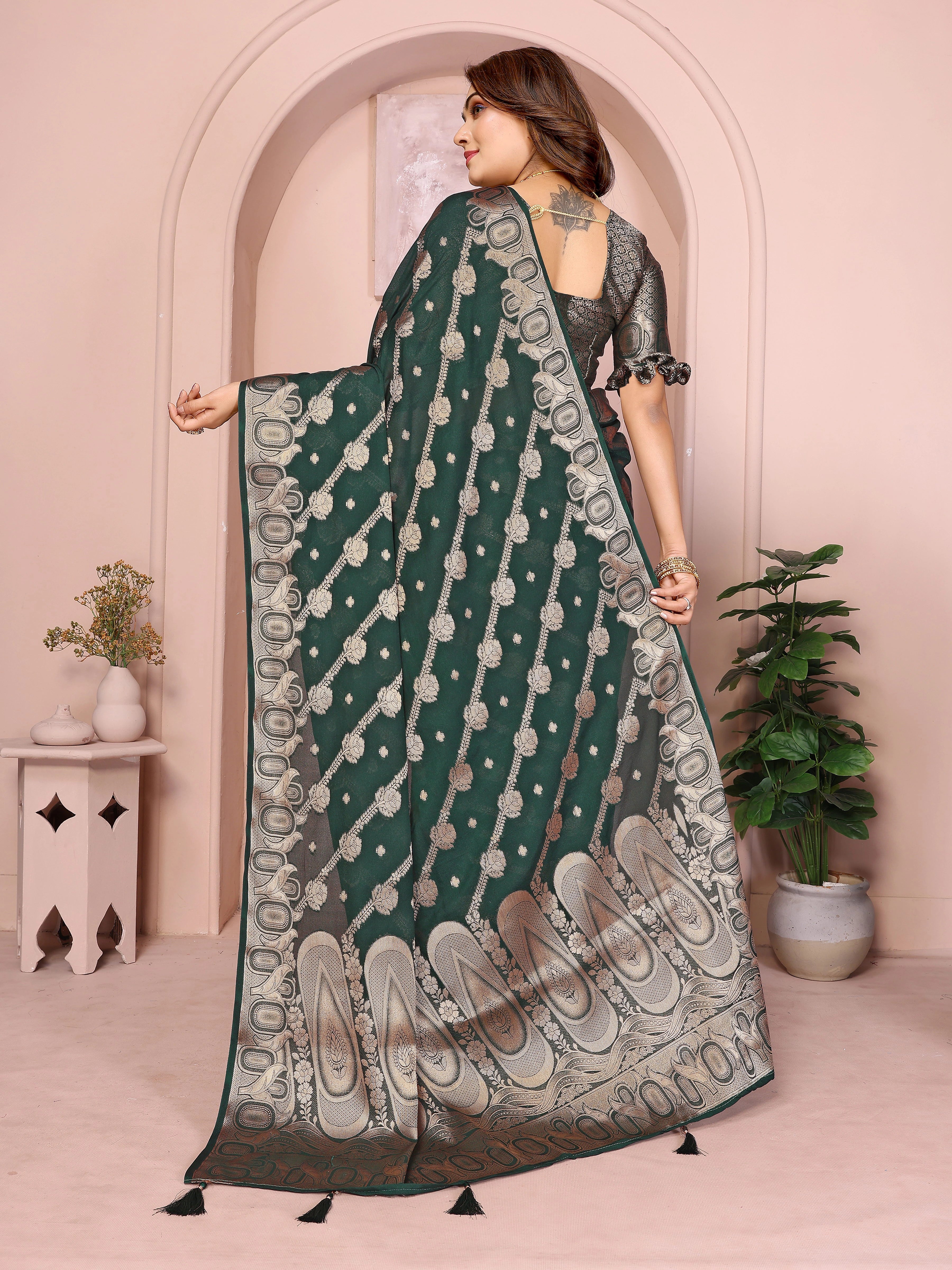 Woman wearing a green saree with a pattern, standing in a room with plants and a pink wall.