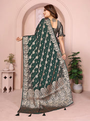 Woman wearing a green saree with a pattern, standing in a room with plants and a pink wall.