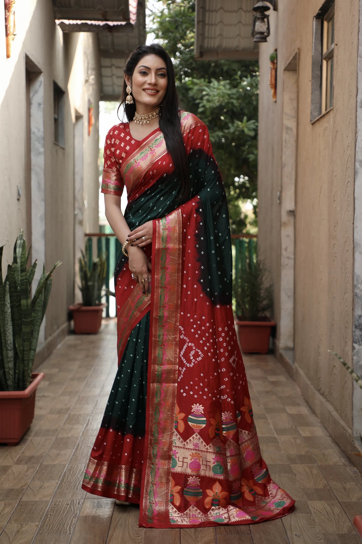 Woman in a red and black saree standing in an outdoor setting with plants and buildings.