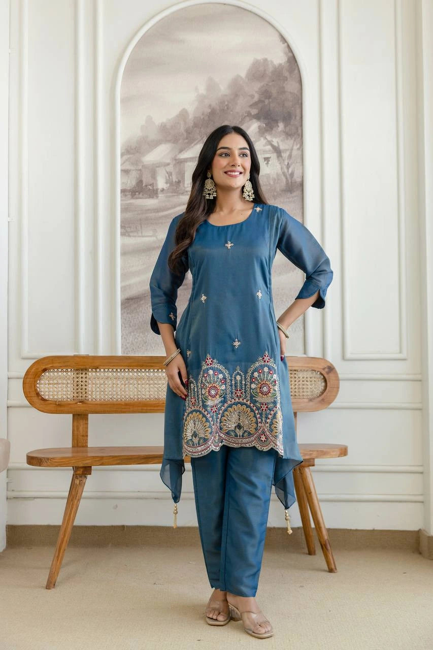 Woman in a blue traditional outfit standing in a room with a wooden bench and decorative wall.