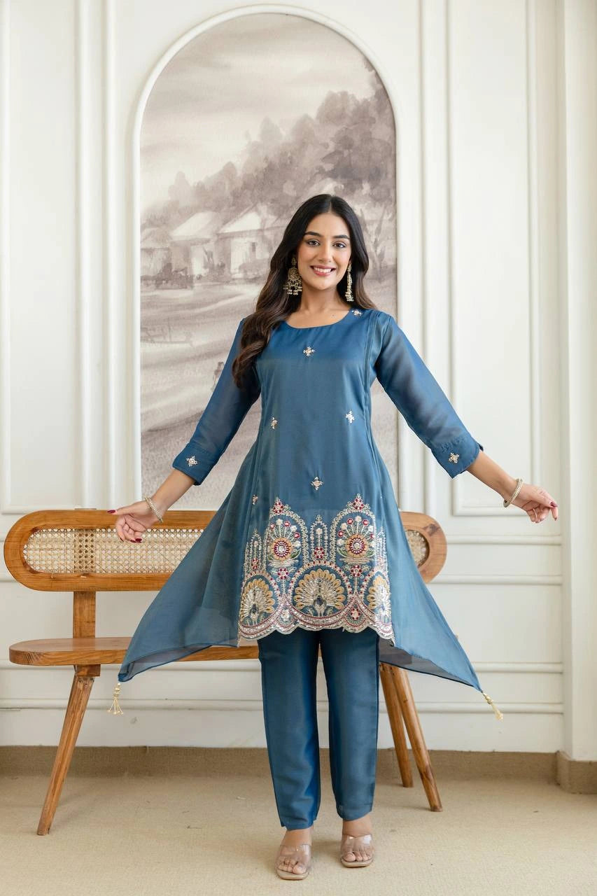 Woman wearing a blue embroidered top and pants standing in a room with a wooden bench and wall art.