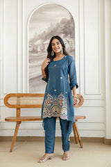 Woman in a blue traditional outfit standing in a room with a wooden bench and decorative wall.