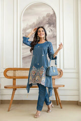 Woman in a blue traditional outfit standing next to a wooden bench with a decorative wall in the background.