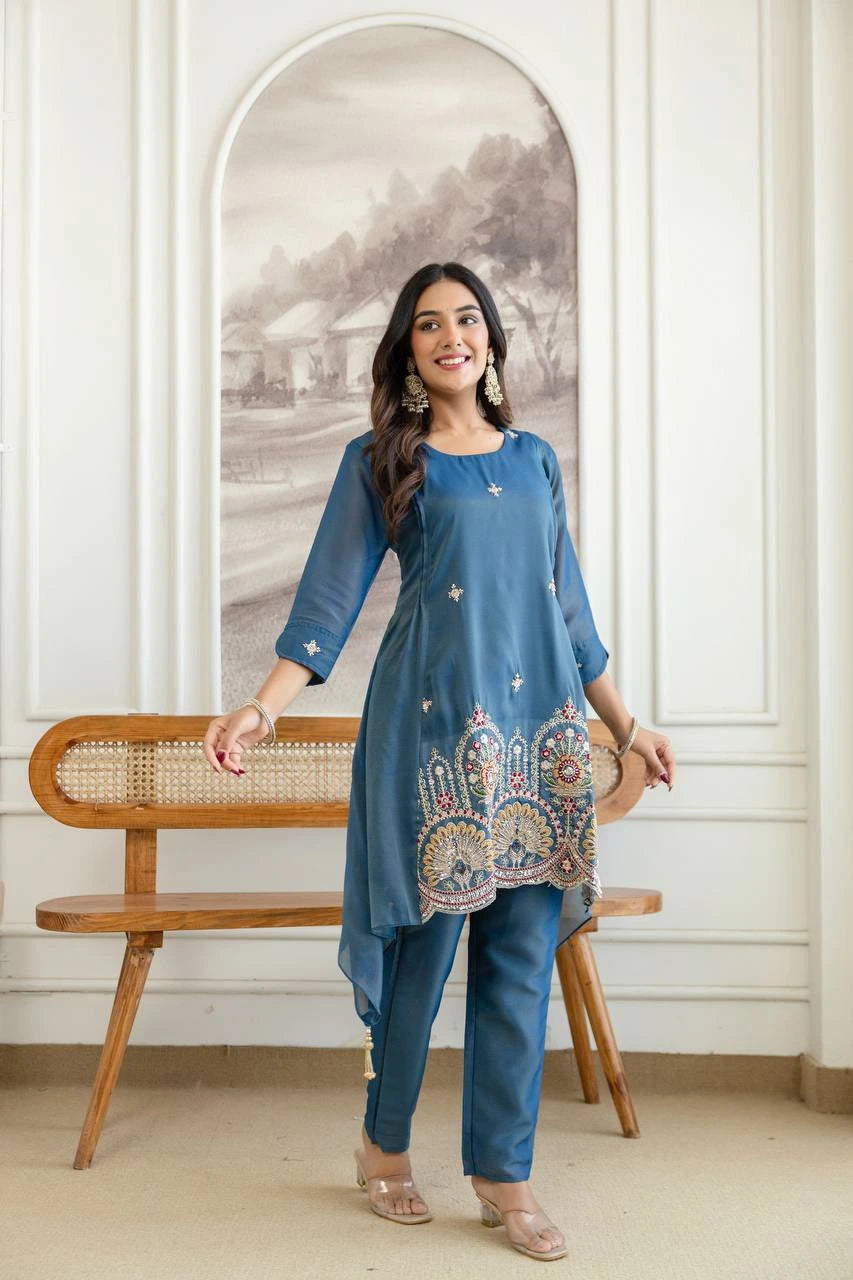 Woman in a blue traditional outfit with embroidery, standing in a room with a wooden bench and wall art.