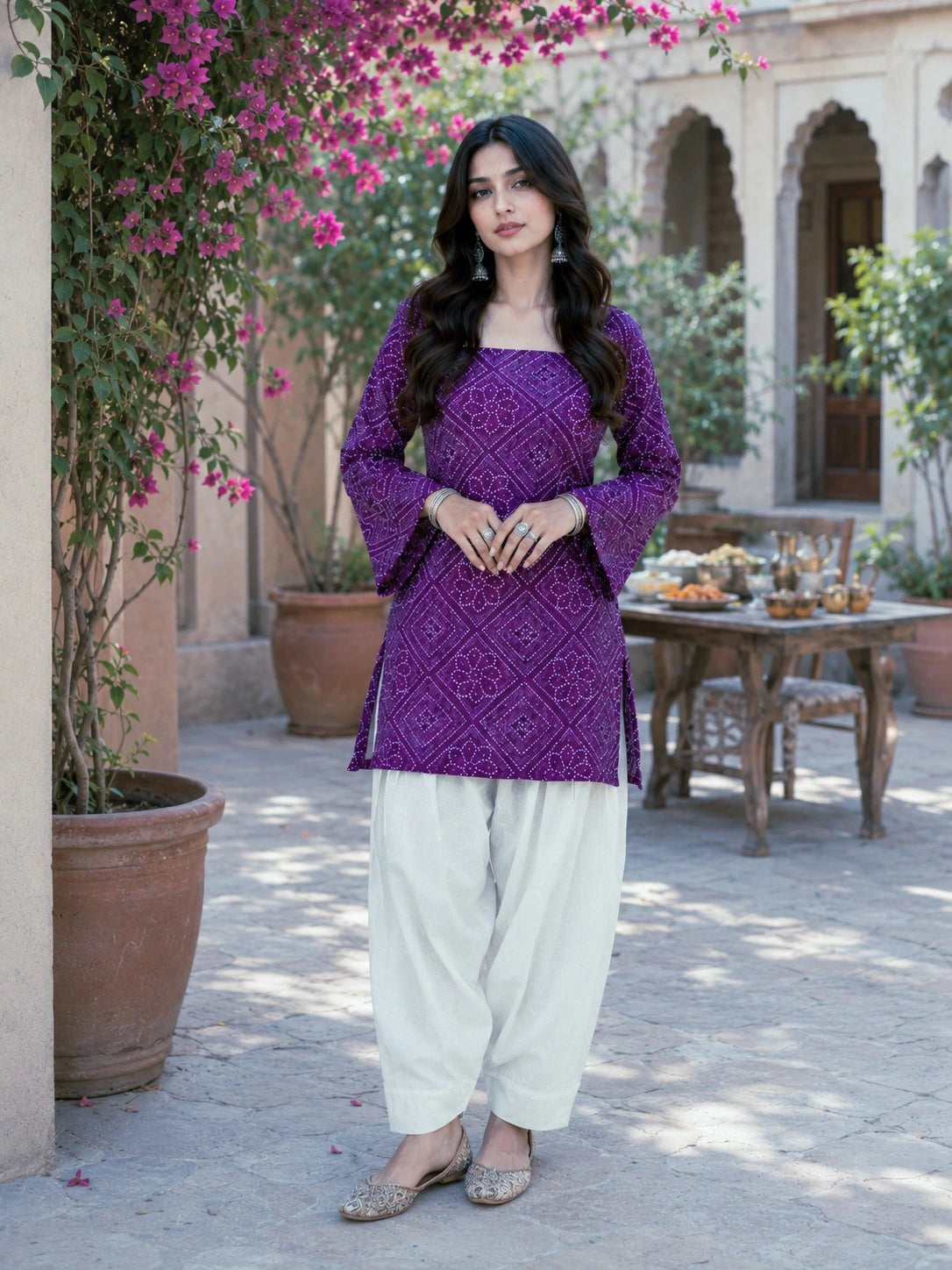 Woman in a purple top and white pants standing in a courtyard with flowers and a table in the background.