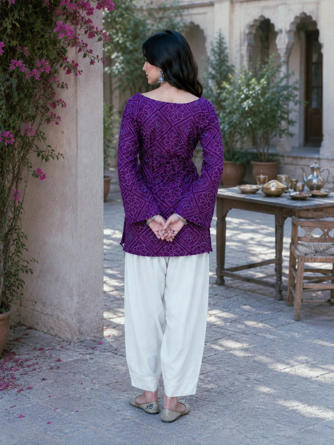 Woman in a purple top and white pants standing in an outdoor setting with plants and a table.