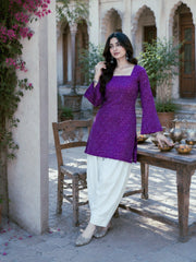 Woman in a purple top and white pants standing in an outdoor setting with a table and chairs.