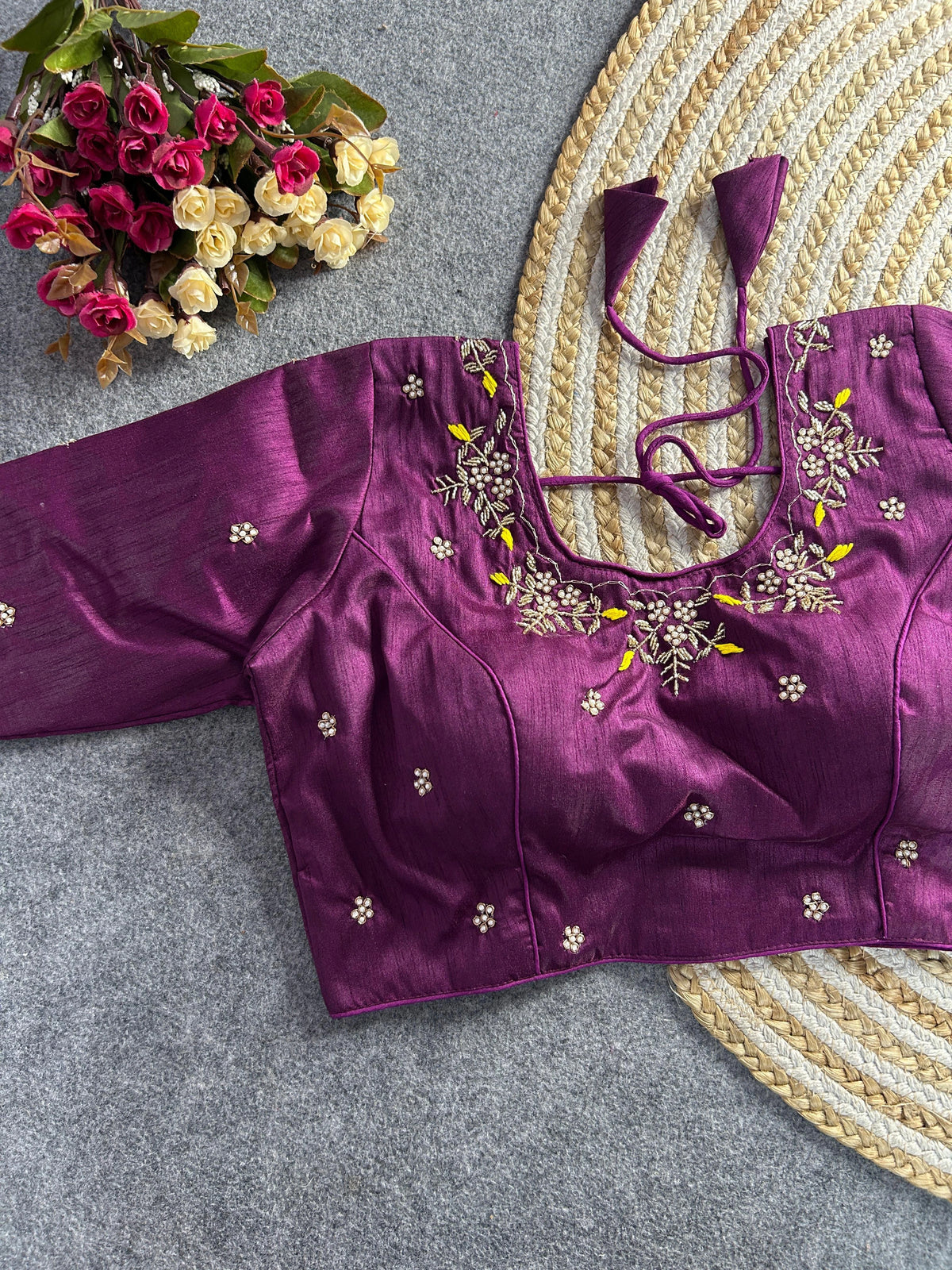 The front of the plum silk blouse showcases a curved V-neckline with the same detailed pearl and gold floral embroidery and yellow accents. The garment is tailored with structured princess-line seams and is adorned with scattered embroidered pearl motifs. The blouse is laid flat on a grey textured background next to a round jute mat and a bouquet of pink and white flowers.
