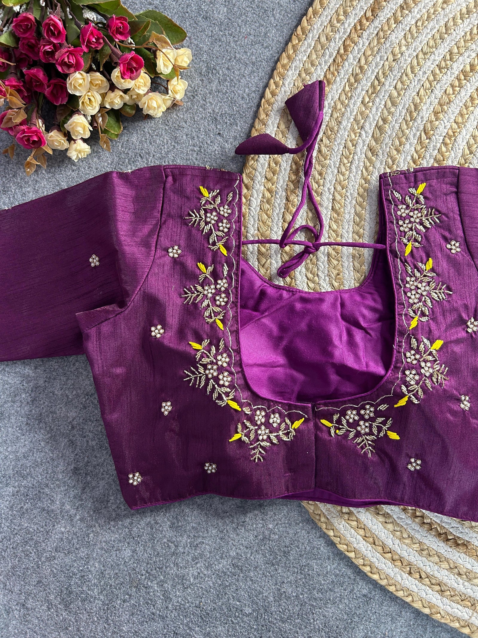 A deep plum-colored raw silk blouse featuring a wide U-shaped back neckline. The neckline is bordered with intricate hand-embroidery of gold and pearl floral sprigs, accented by vibrant lemon-yellow threadwork. It includes matching plum tie-back cords with triangular tassels, and small pearl floral buttis are scattered across the back and sleeves.