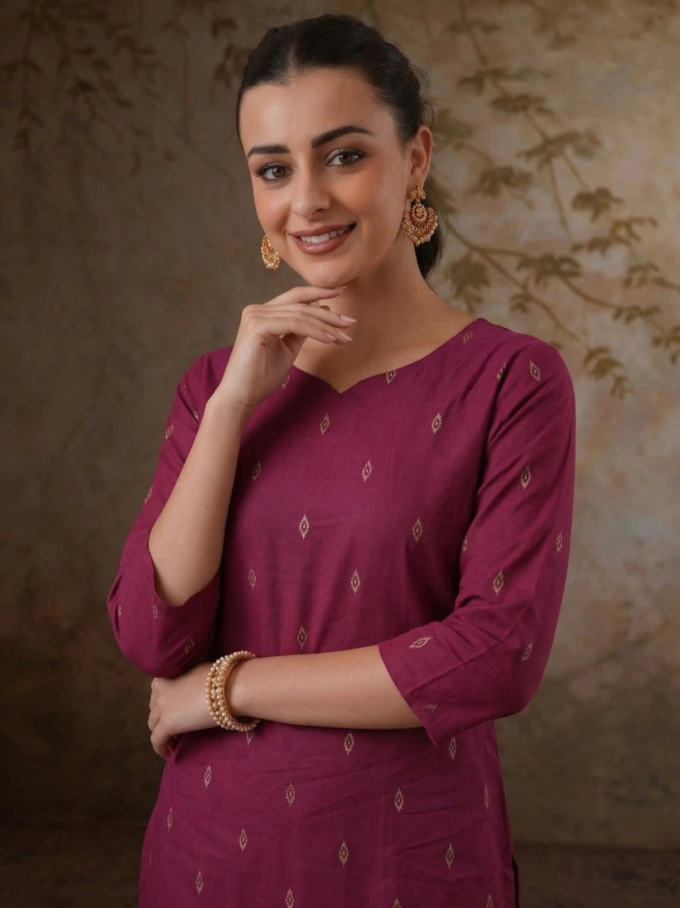 Woman wearing a maroon traditional outfit with gold jewelry against a textured wall.
