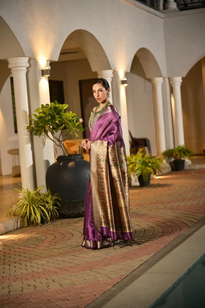 Woman wearing a deep purple silk saree with a rich gold woven pallu, posing in a warm-lit courtyard with arches and greenery, giving an elegant and traditional festive vibe.
