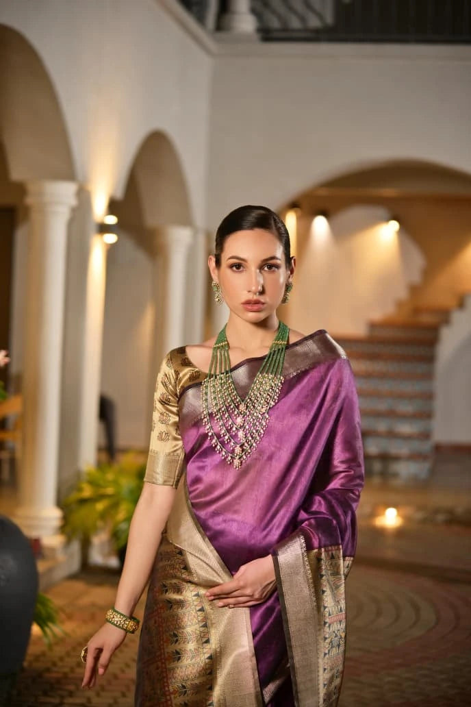A woman stands gracefully in an arched courtyard, wearing a shimmering purple silk saree with intricate gold borders and embroidery. She accessorizes with multi-layered green beaded necklace, matching bangles, and sleek hair.
