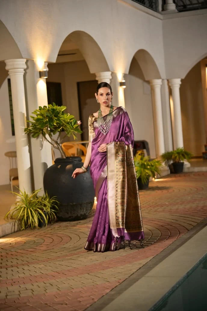 Elegant woman in a rich purple silk saree with gold border, standing in a softly lit courtyard with arches, plants, and warm ambient lighting, exuding a graceful and festive look.