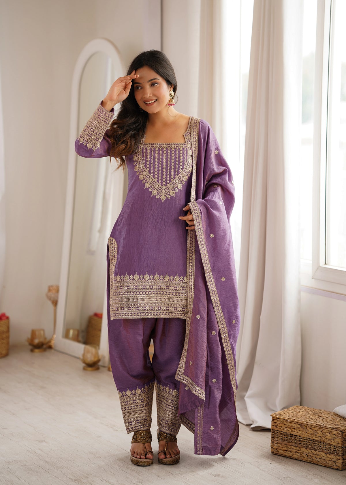 Woman in a purple traditional outfit with intricate patterns standing in a room with white curtains.