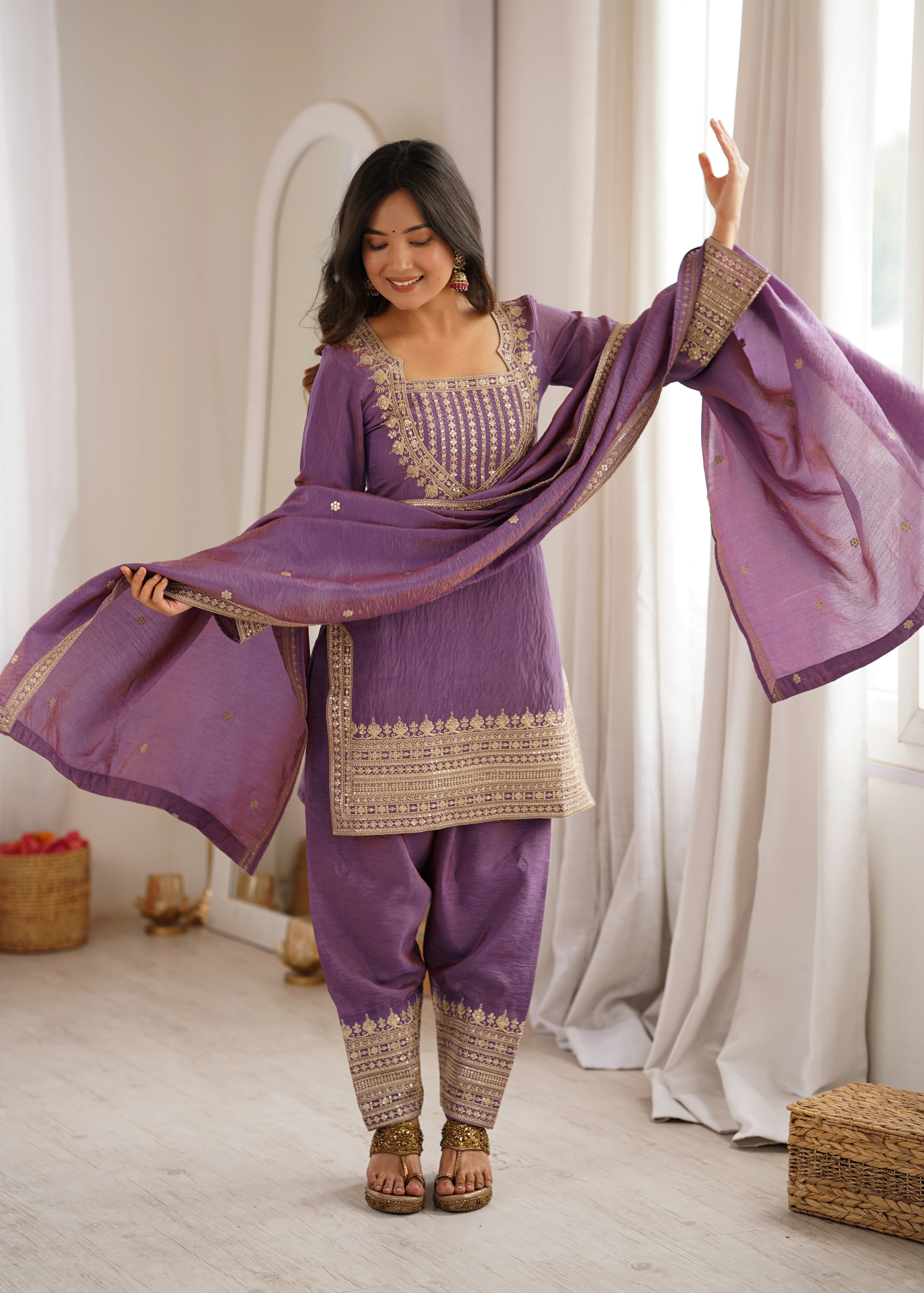 Woman in a traditional purple outfit with gold embroidery standing in a room with white curtains.