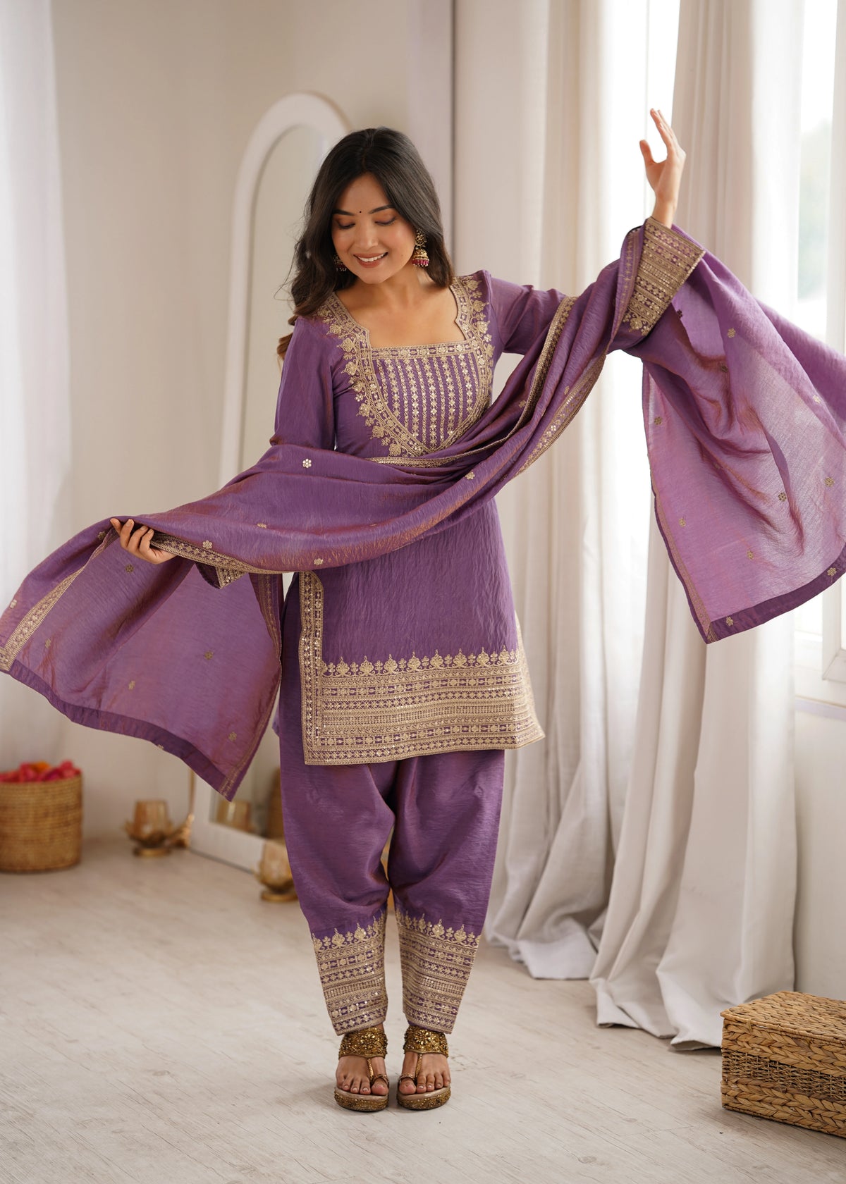 Woman in a traditional purple outfit with gold embroidery standing in a room with white curtains.