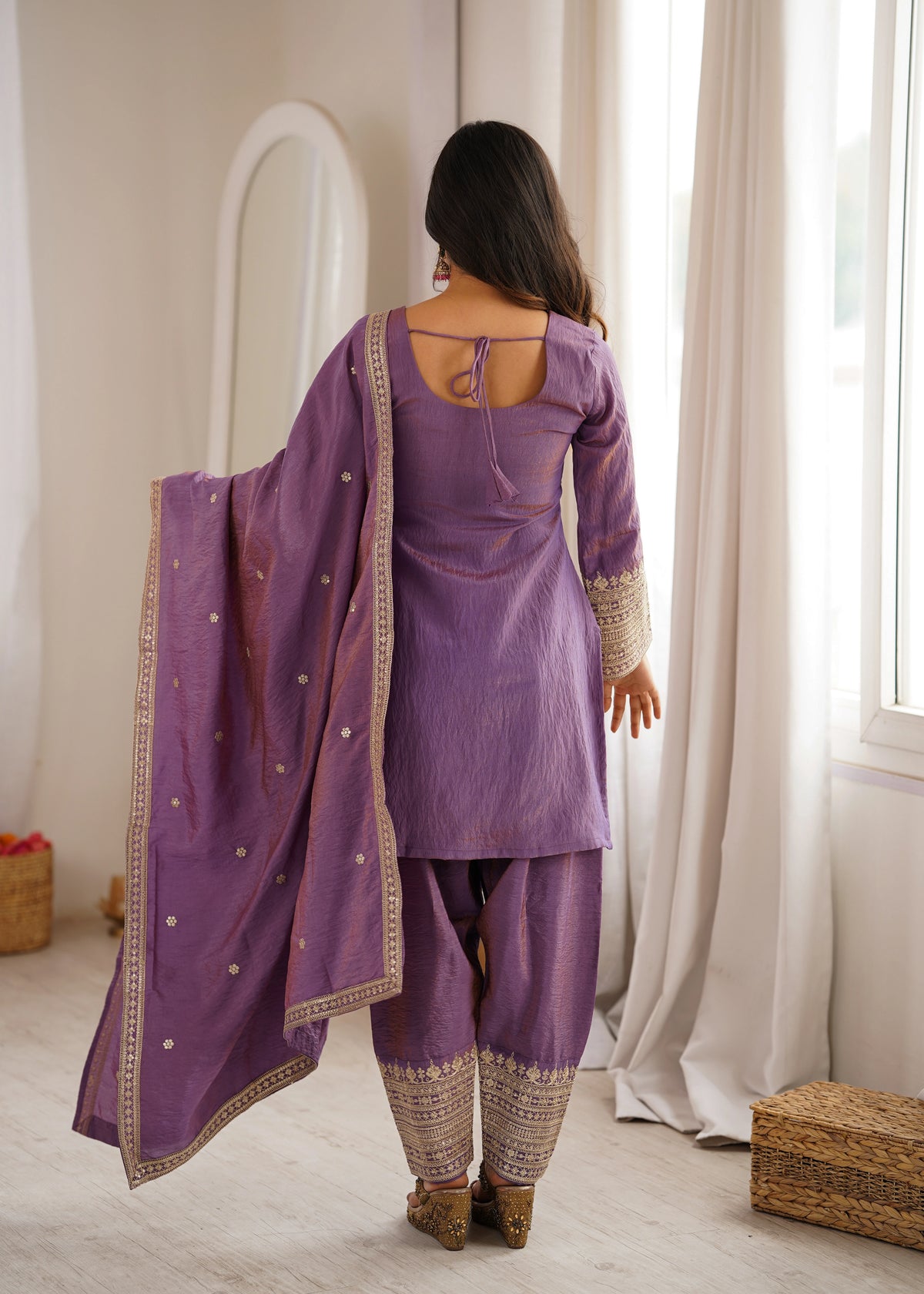 Woman in a purple traditional outfit standing in a room with light-colored walls and curtains.