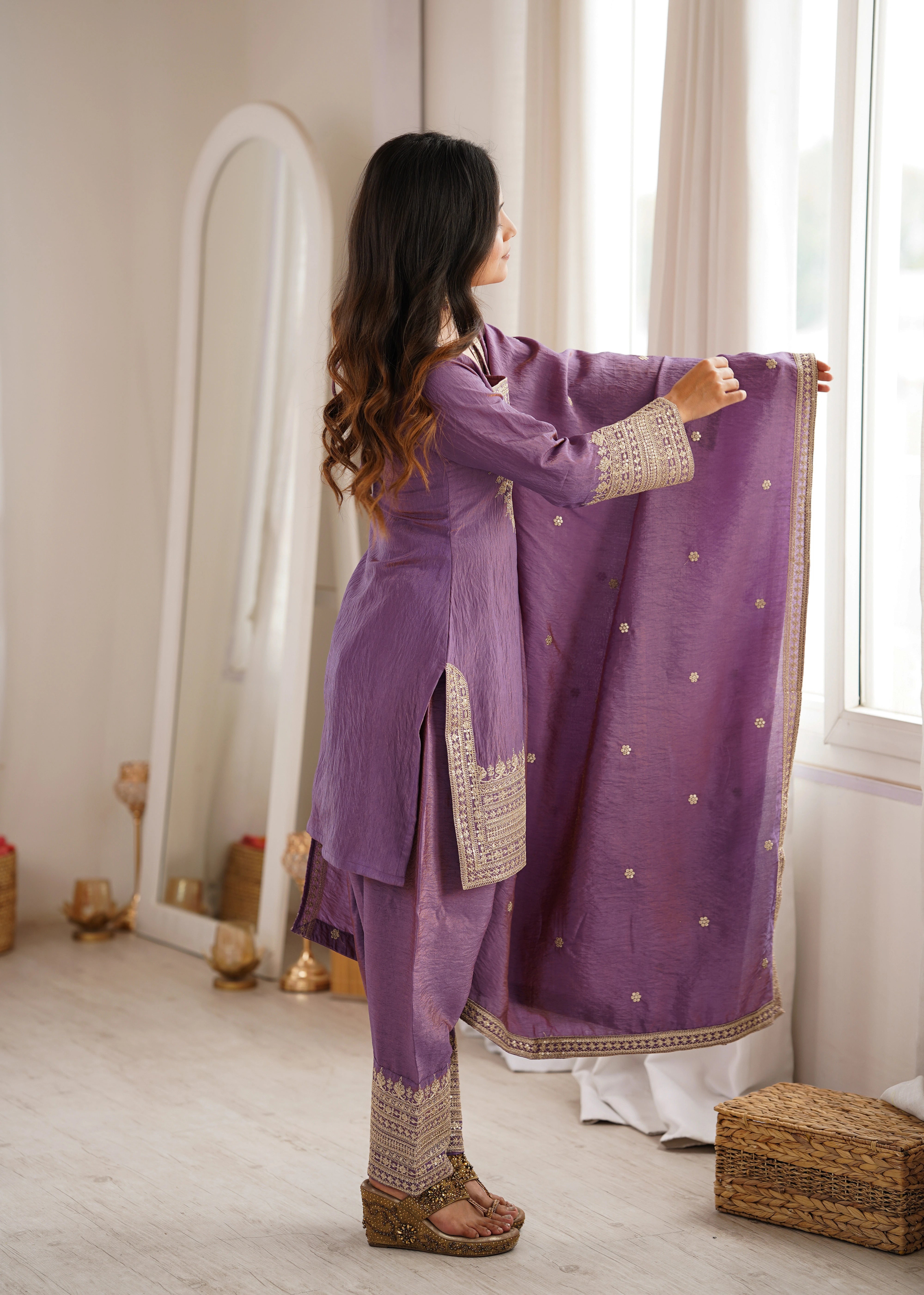 Woman in a purple traditional outfit standing by a window in a well-lit room.