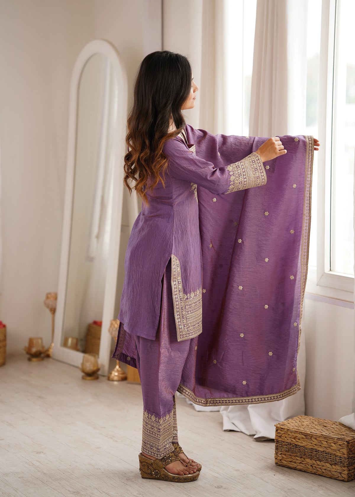 Woman in a purple traditional outfit standing by a window in a well-lit room.