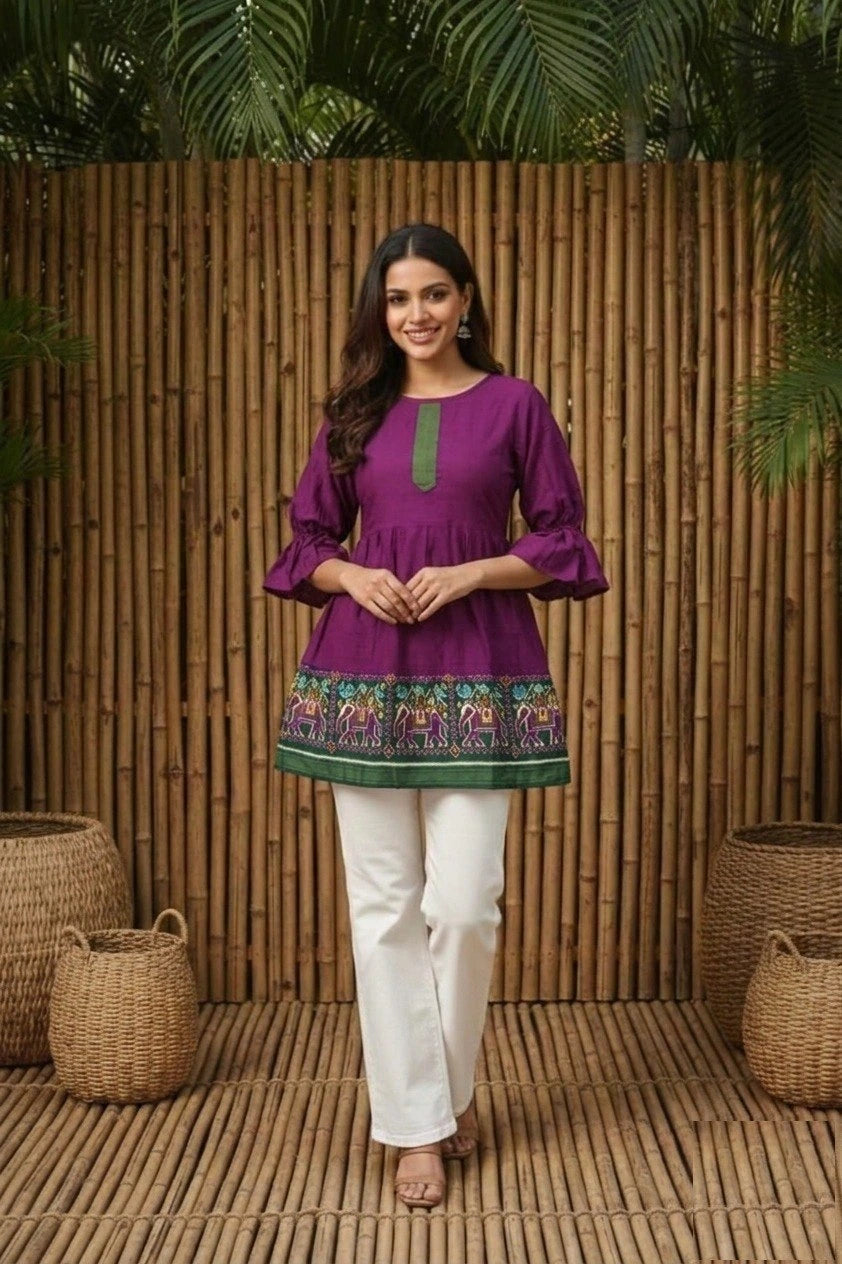 Woman wearing a purple embroidered top and white pants standing against a wooden panel background.