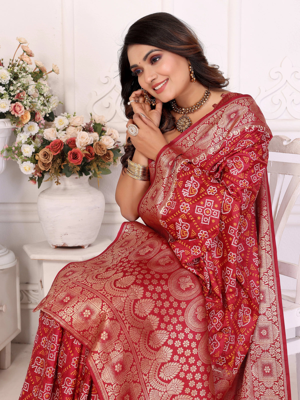 Woman wearing a red saree with white patterns, sitting indoors with flowers in the background.