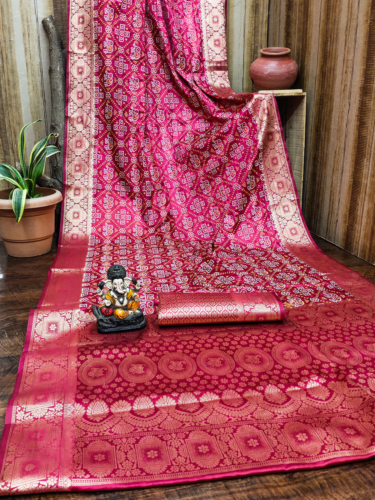 Pink saree with intricate patterns on a wooden surface, with a small statue and plant in the background.