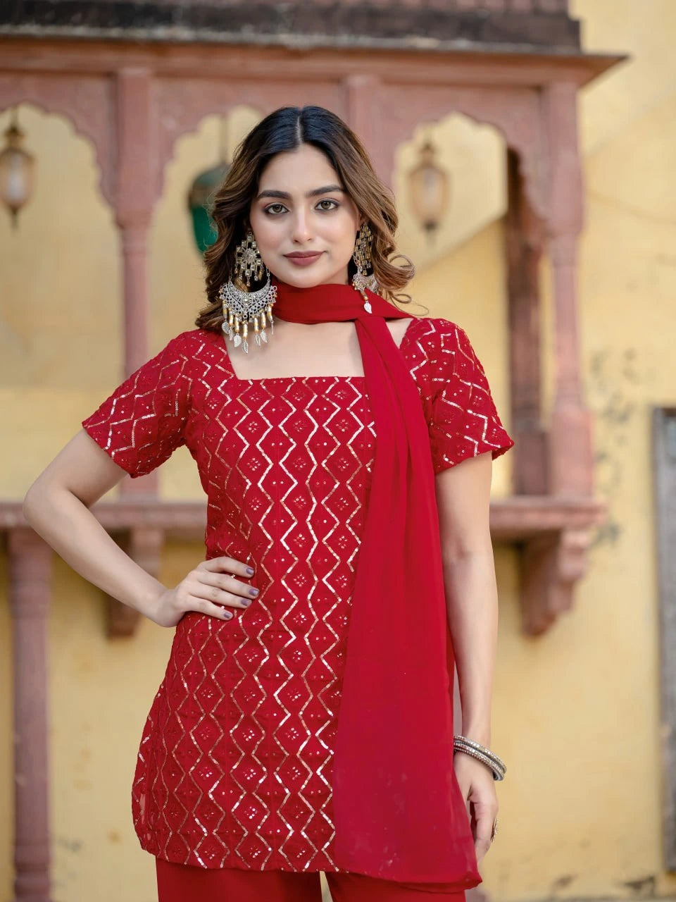 Woman wearing a red patterned outfit with a matching dupatta against a decorative wall.
