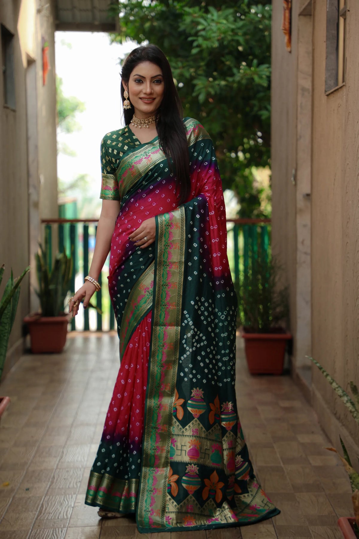 Woman in a colorful saree standing in an outdoor setting with plants and a building.