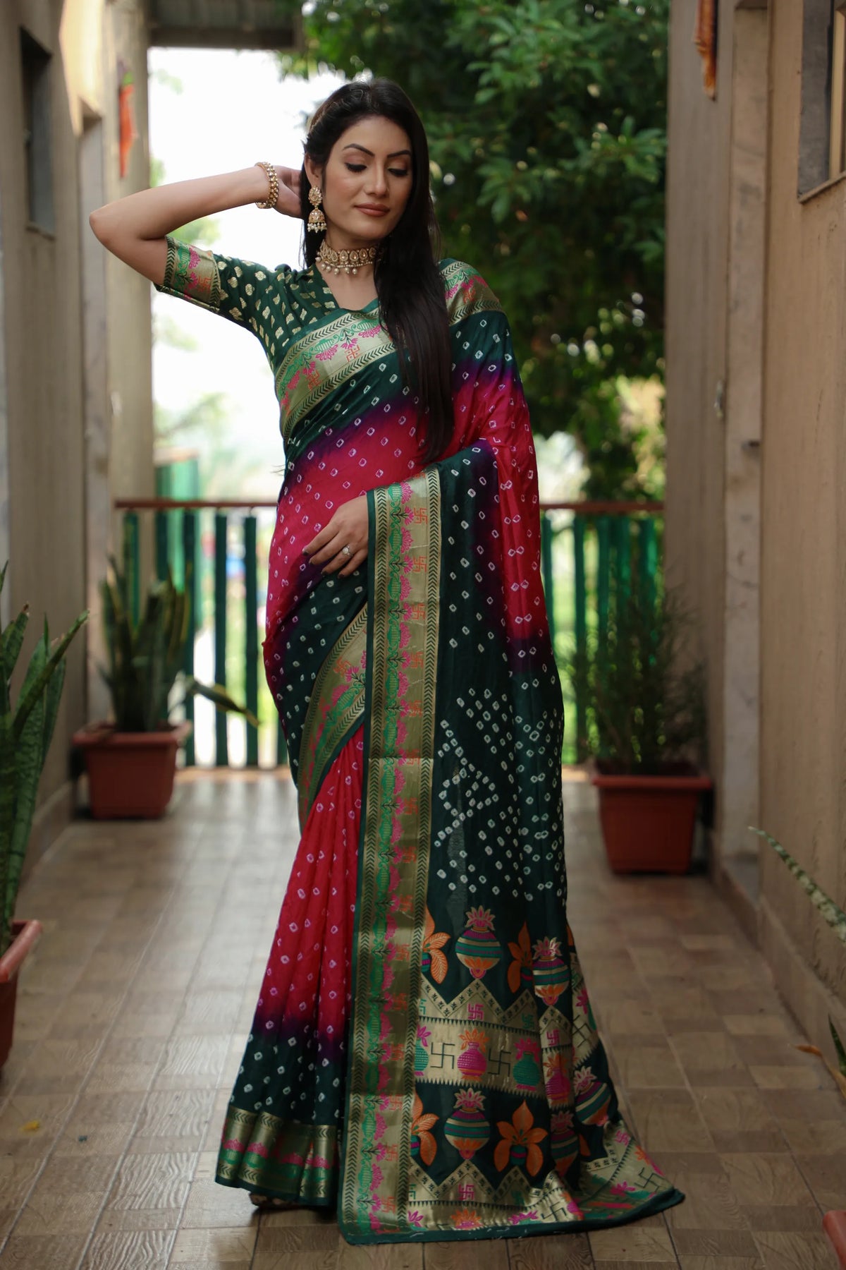 Woman in a colorful saree standing in an outdoor setting with plants and a building.