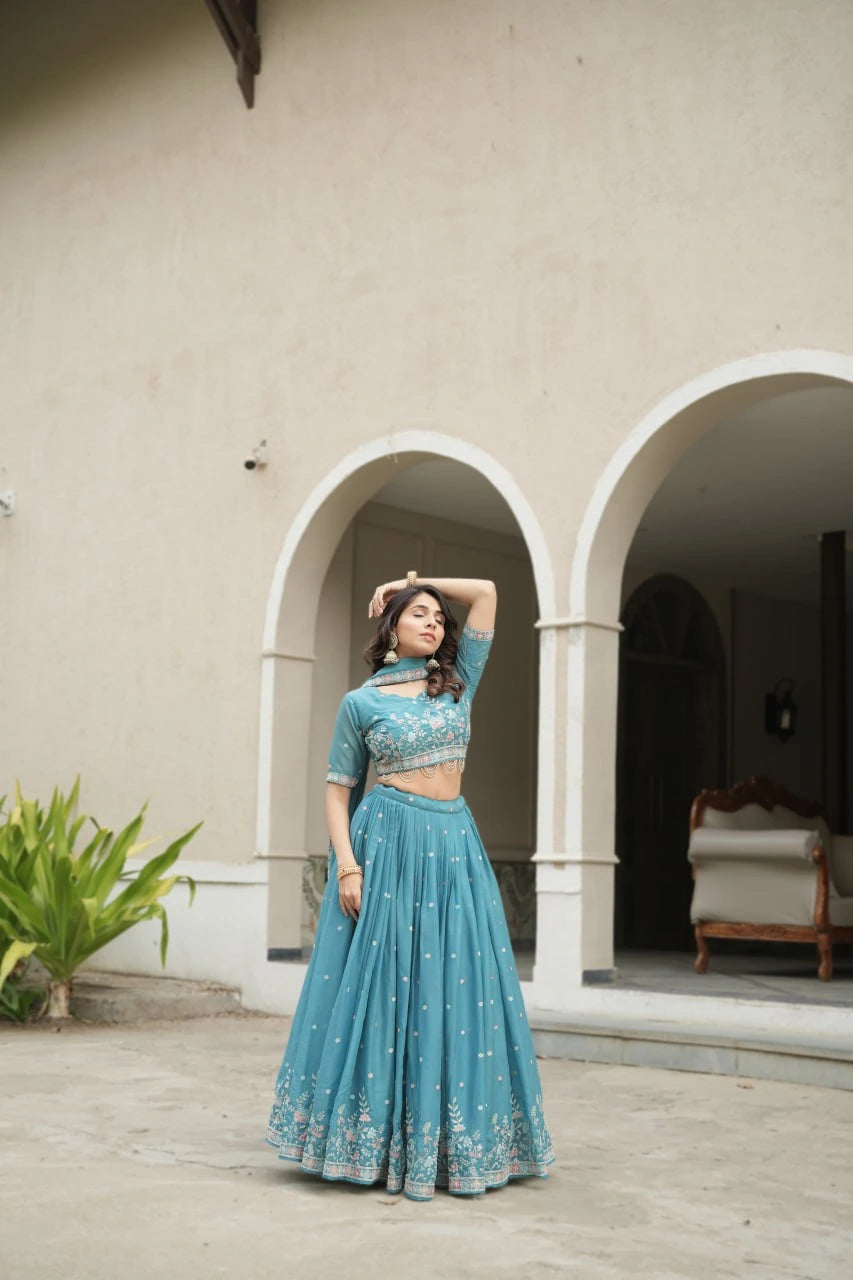 Woman in a teal traditional outfit standing in front of a beige wall with arches.