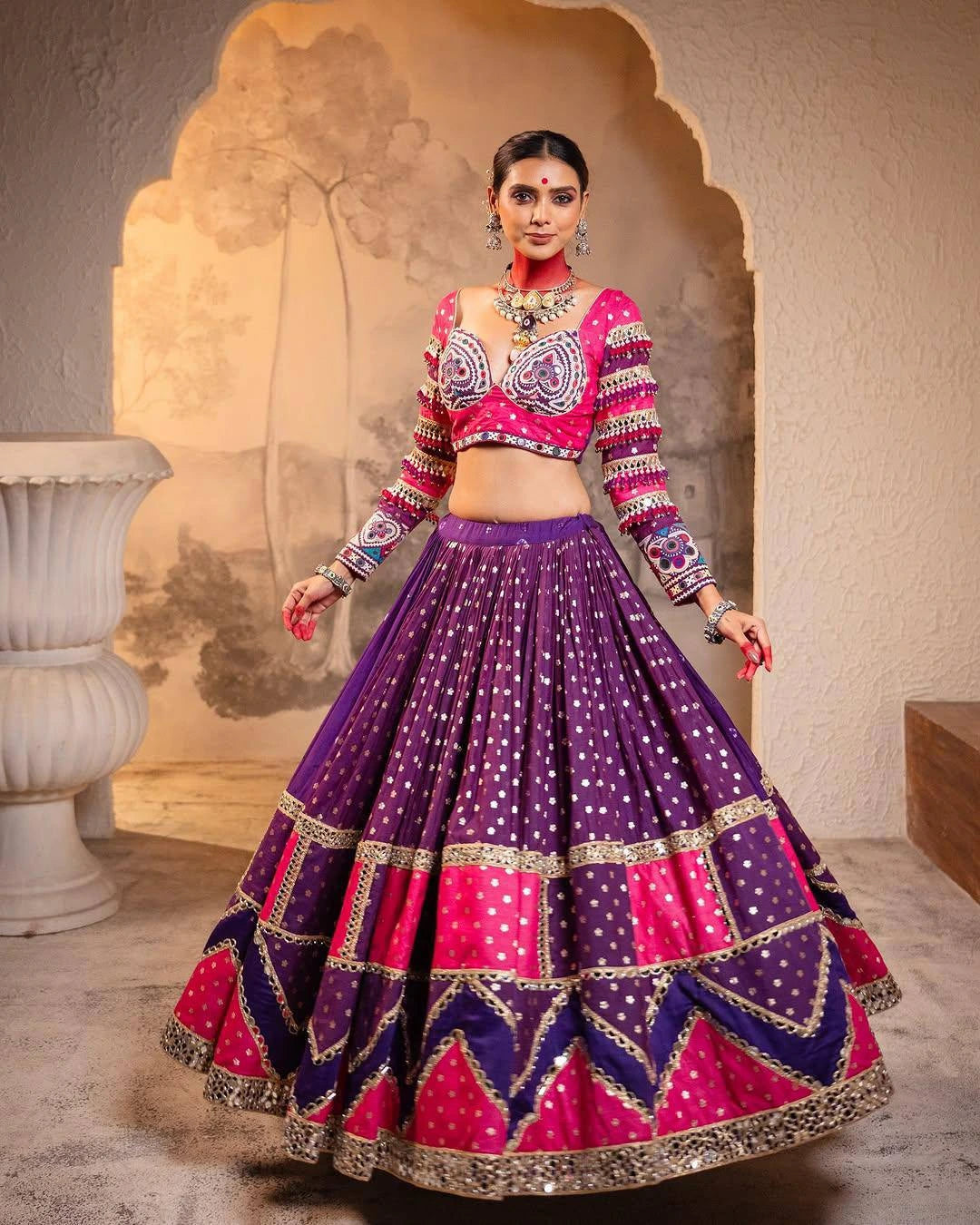 Woman wearing a traditional outfit with a pink blouse and purple skirt in an ornate setting.