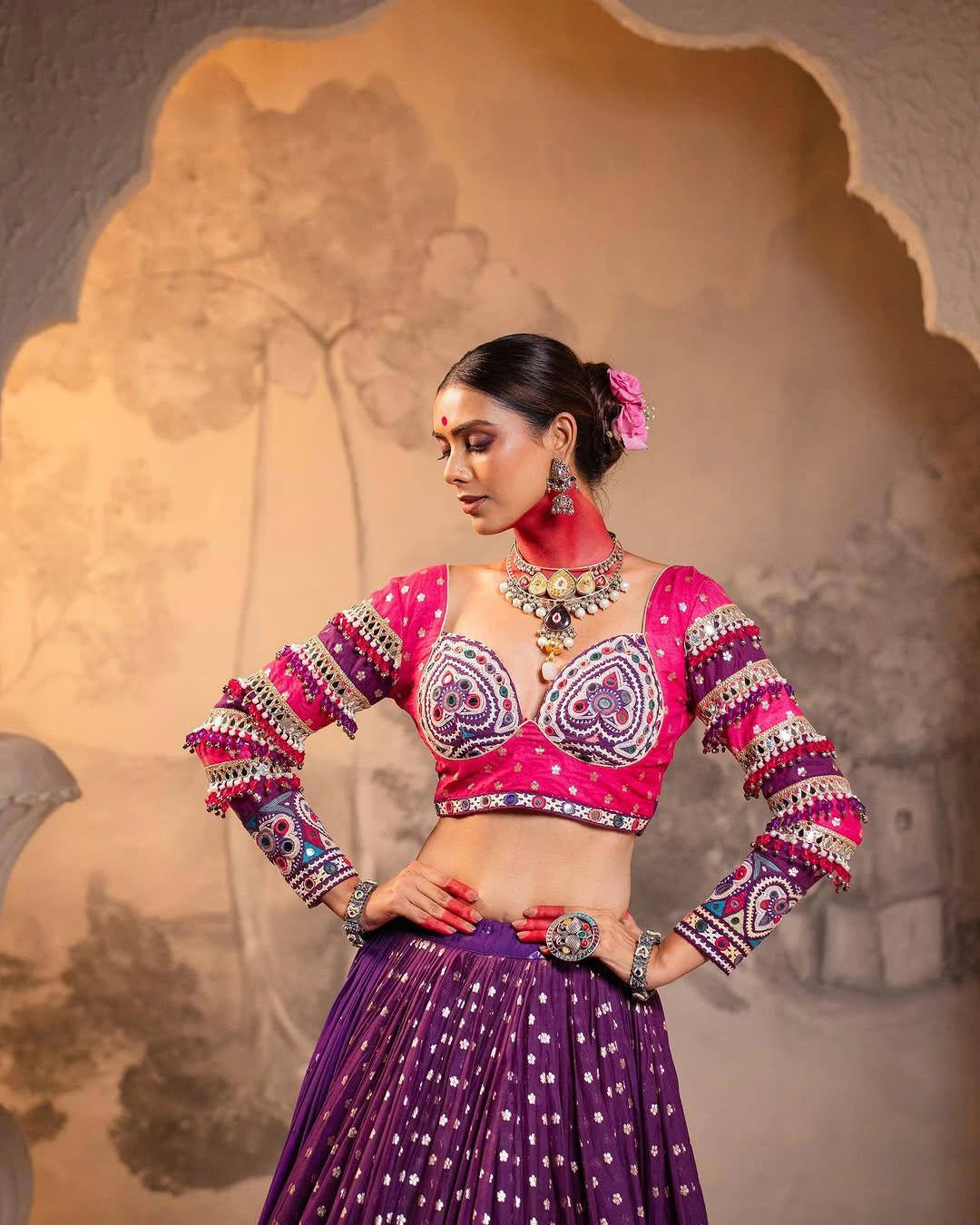 Woman in a vibrant traditional outfit with a pink blouse and purple skirt against a decorative background