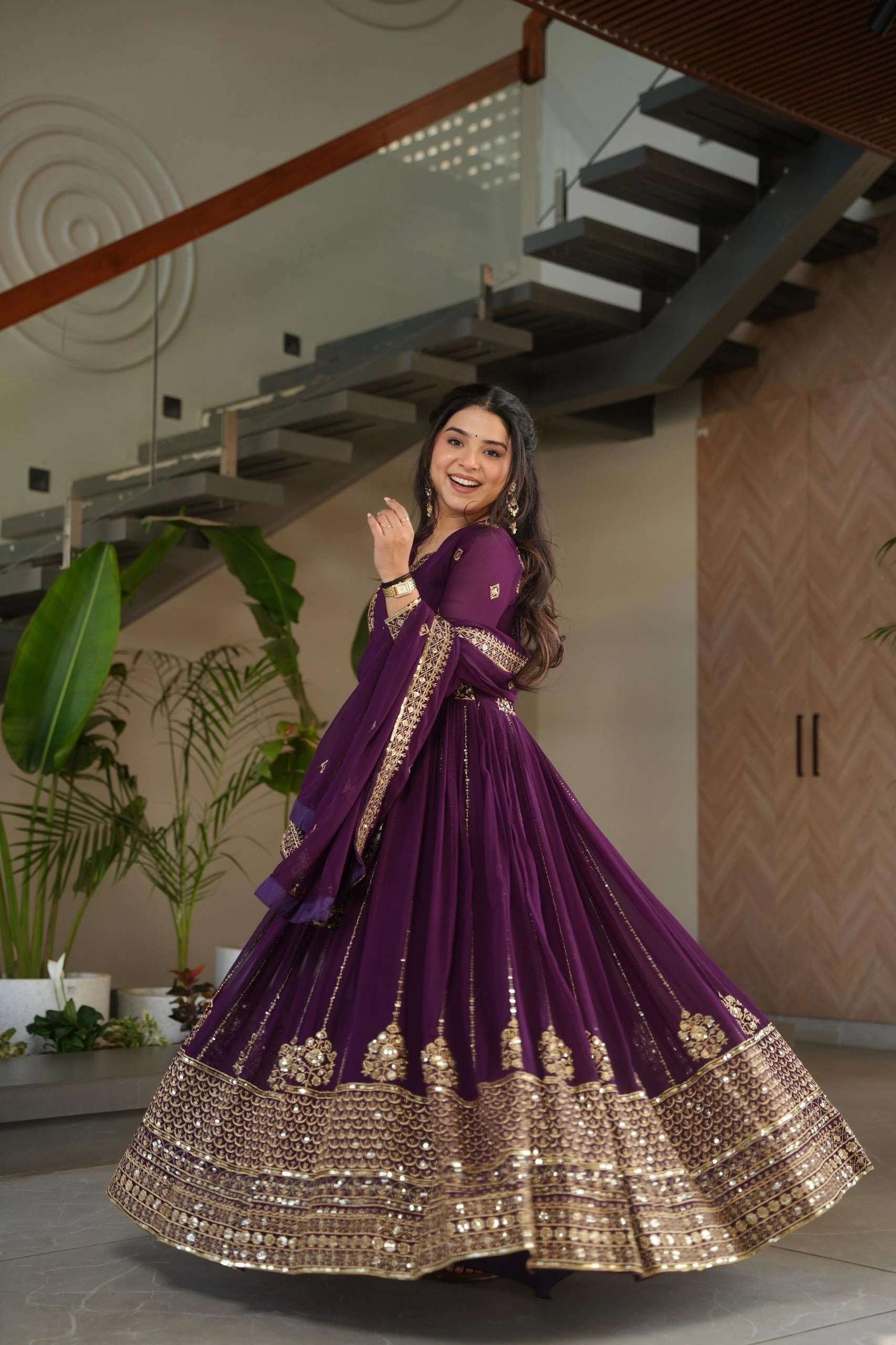 Woman in a purple and gold traditional outfit standing indoors with plants and a staircase in the background.