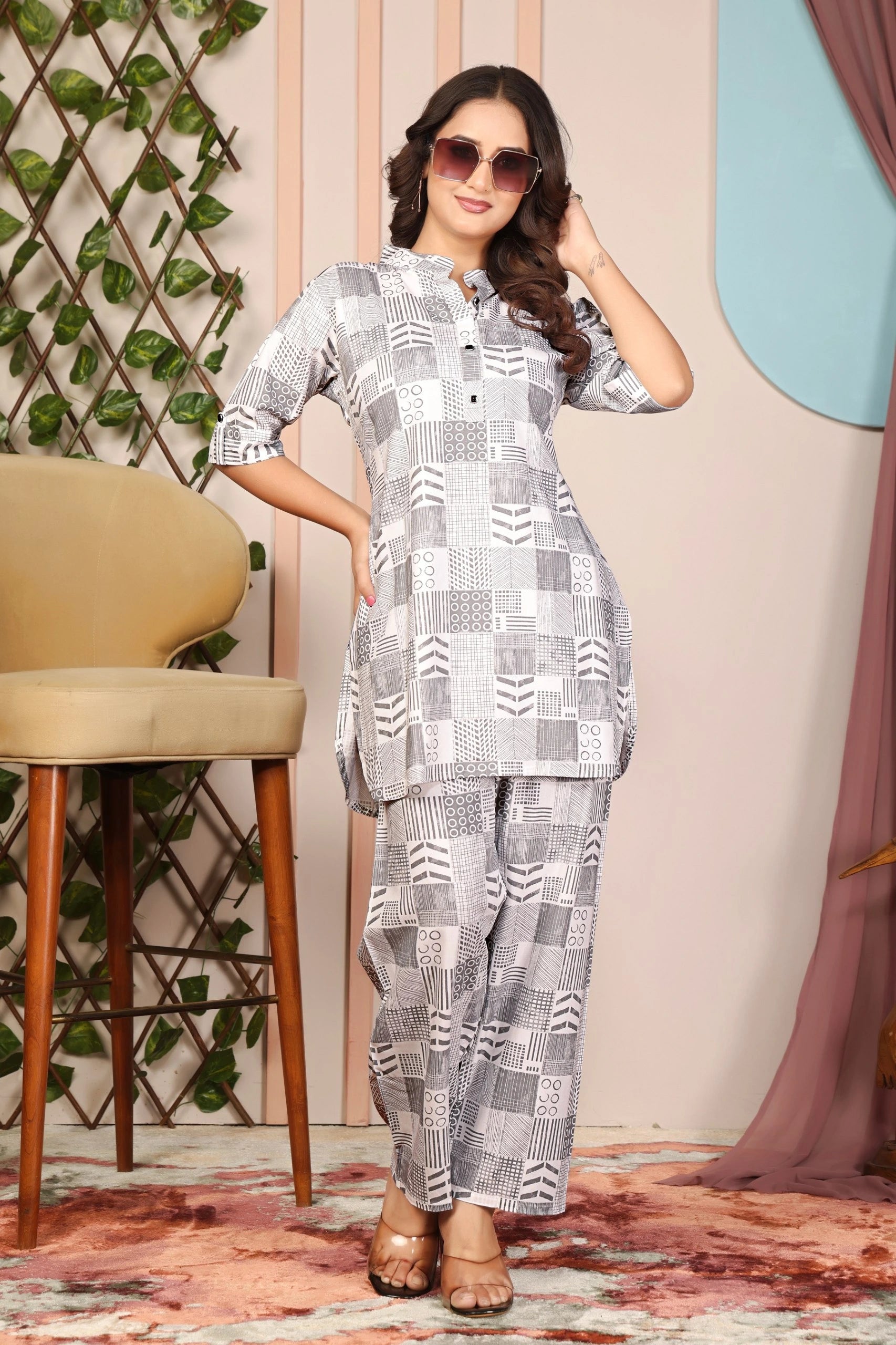 Woman wearing a patterned outfit in a room with a chair and decorative wall.