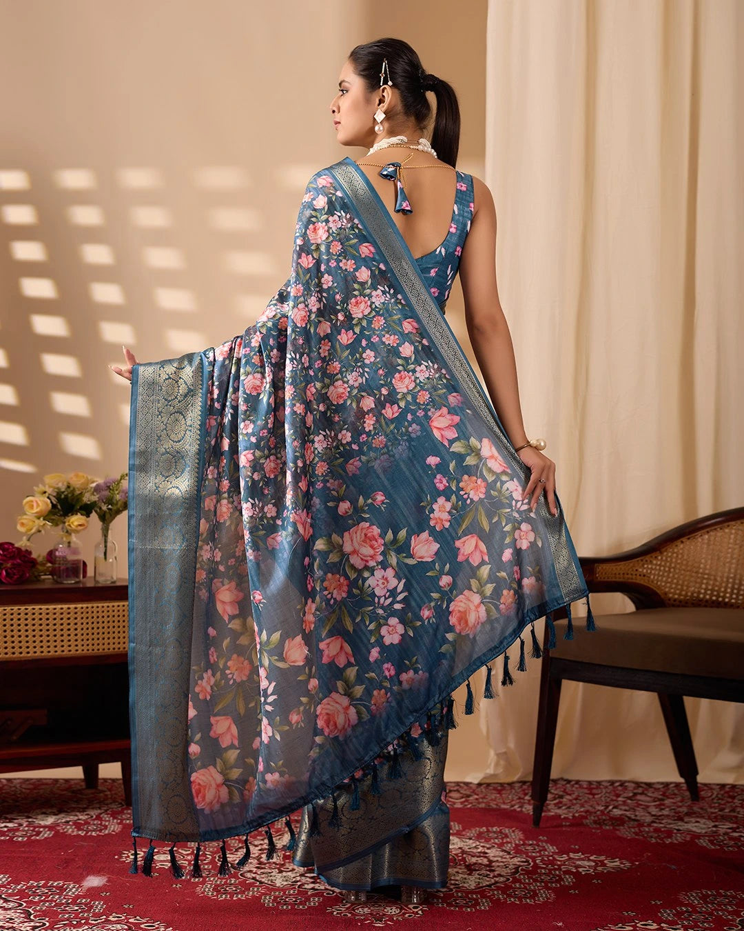 Woman wearing a blue floral saree in a room with a table and chair.