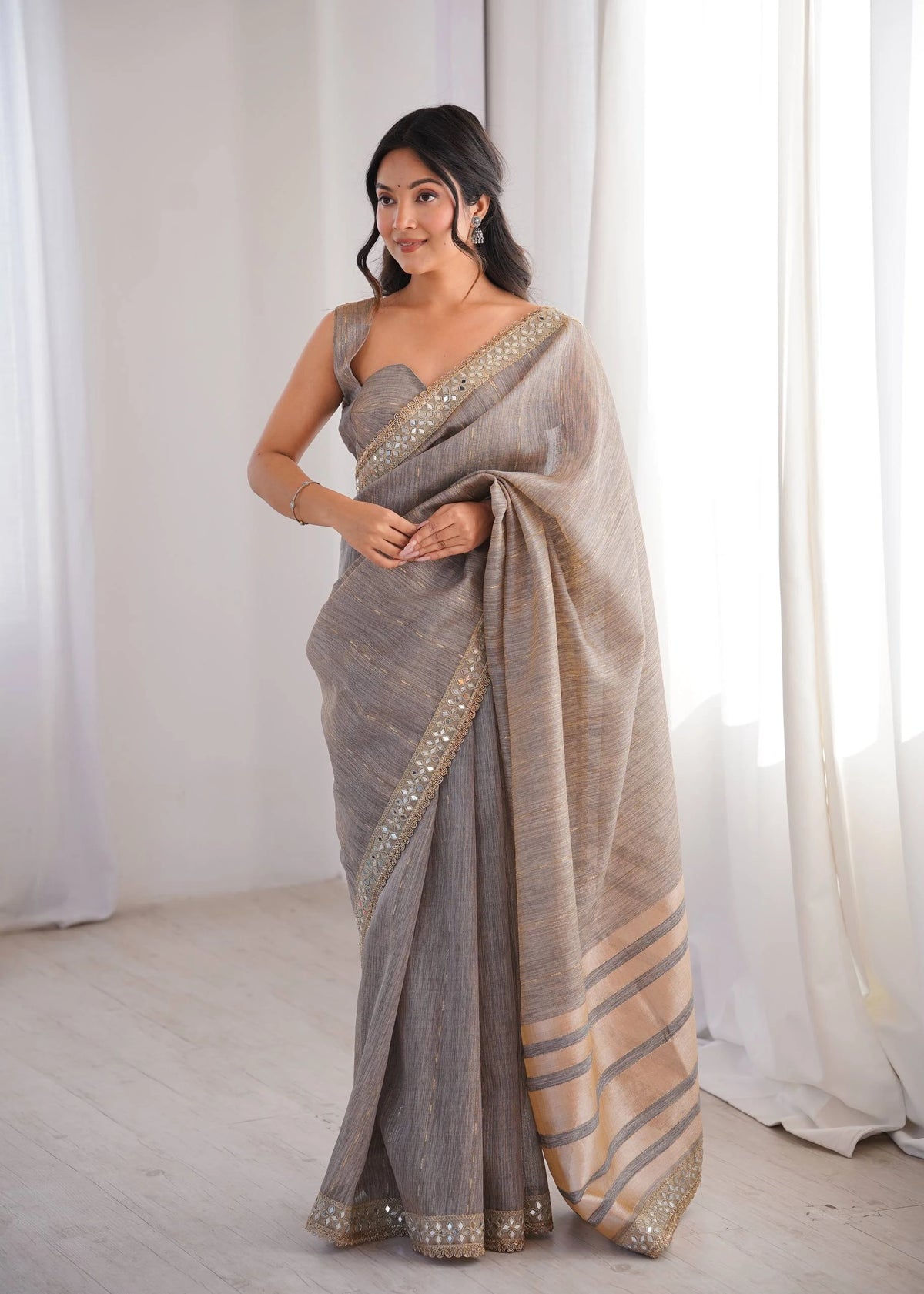 Woman wearing a gray saree with gold accents against a white curtain background