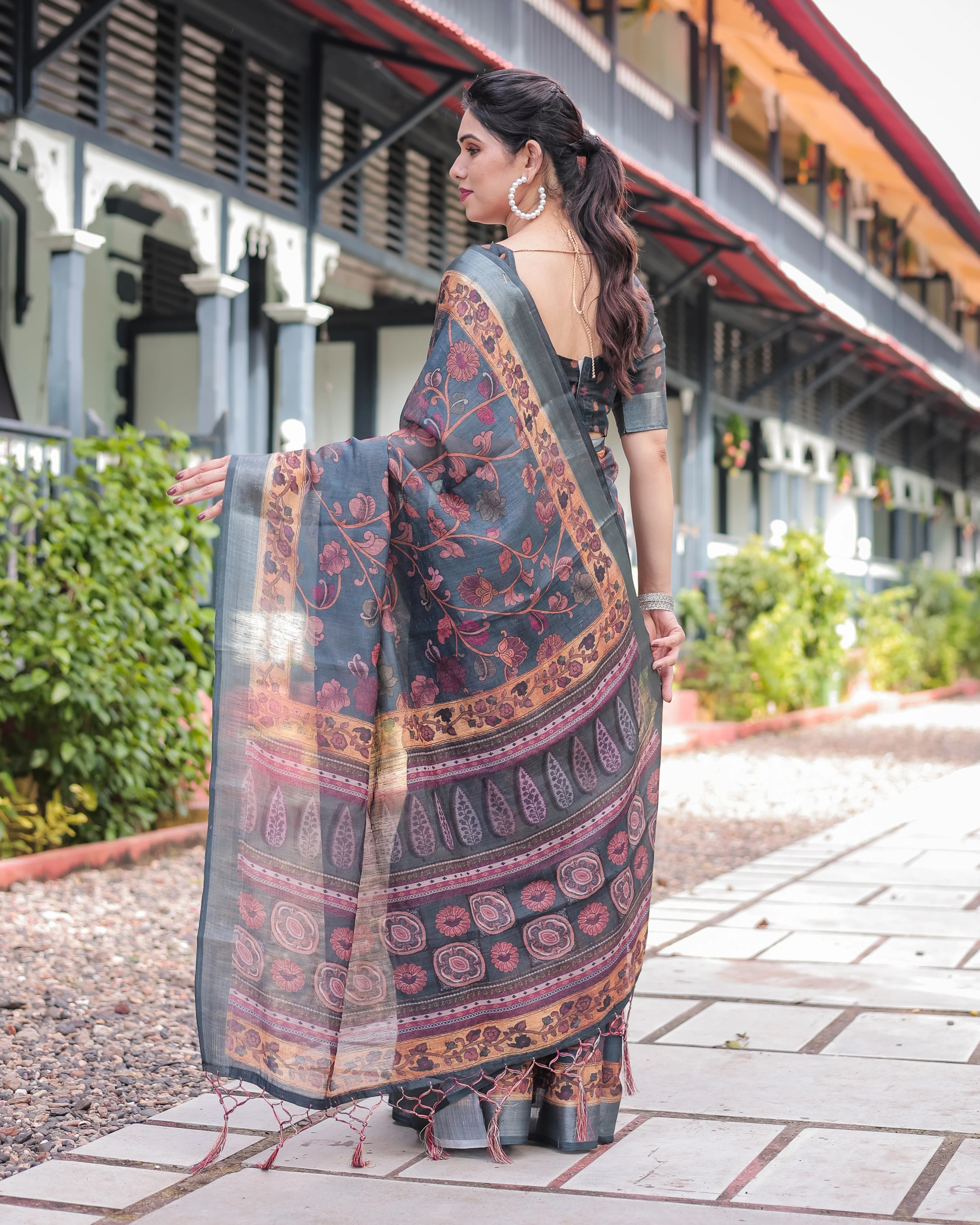 Woman holding a patterned saree on a sidewalk with greenery and a building in the background