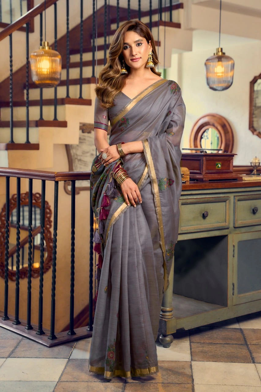 Woman in a gray saree standing in an indoor setting with decorative elements.