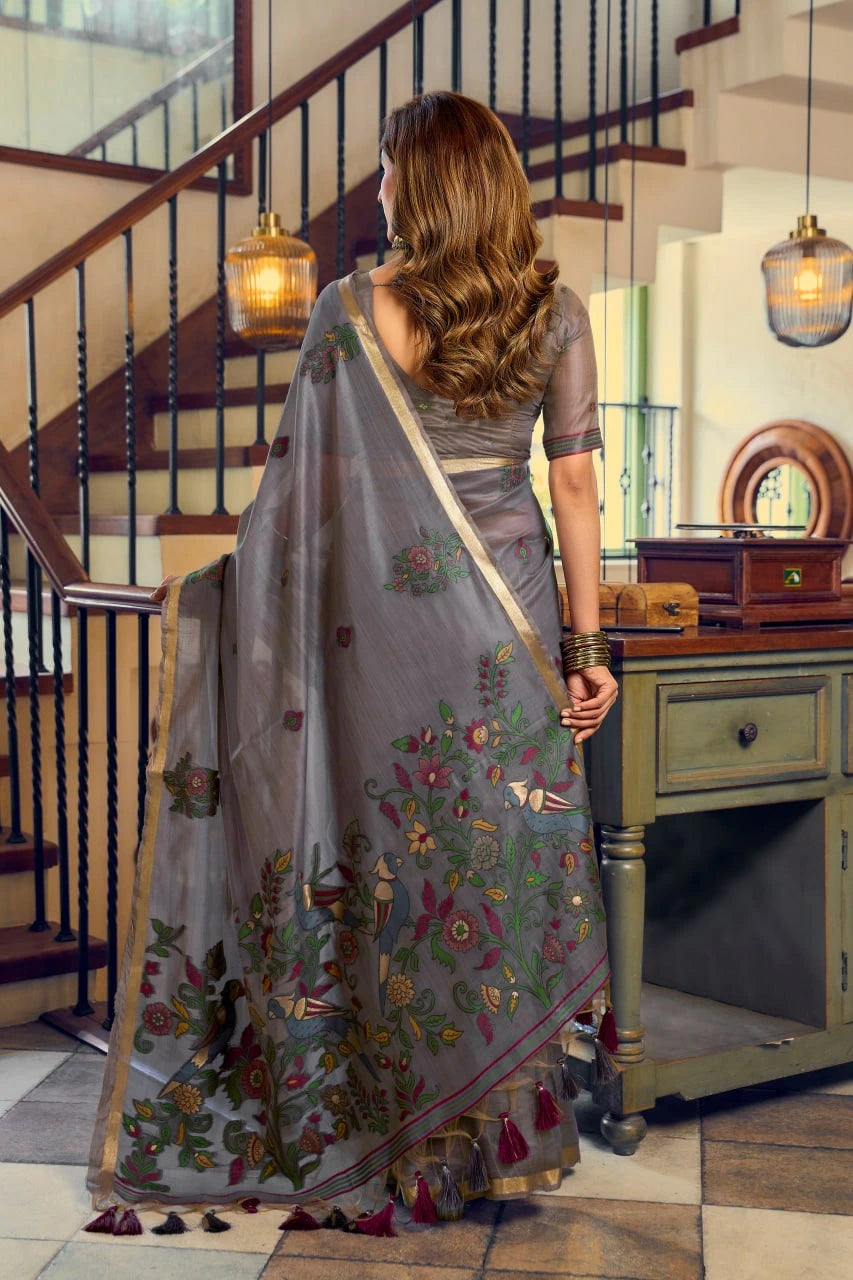 Woman in a gray saree with floral patterns standing in a room with wooden staircase and furniture.