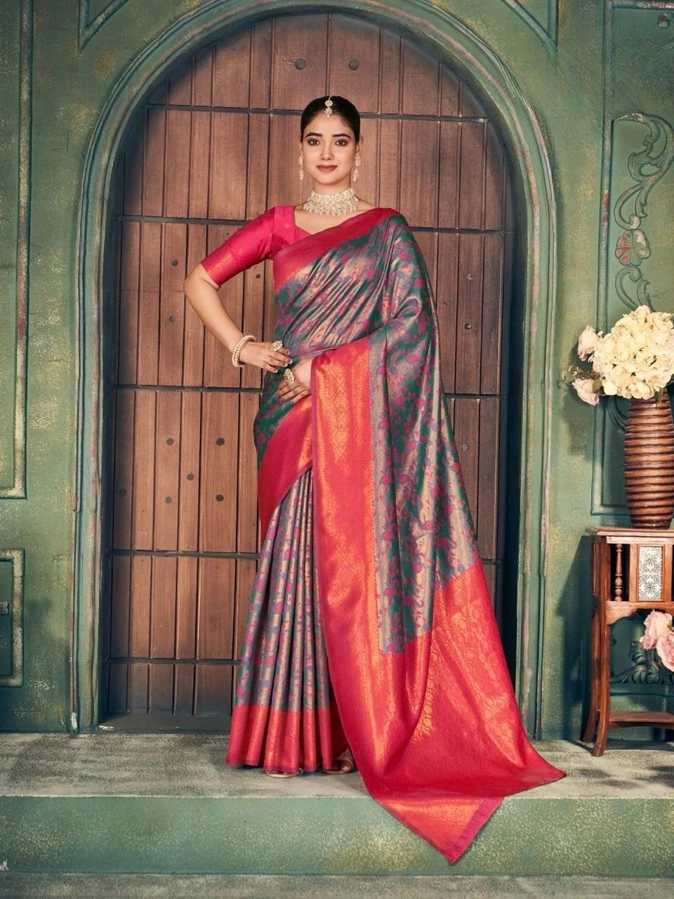 Woman in a red and gray saree standing in front of a wooden door.
