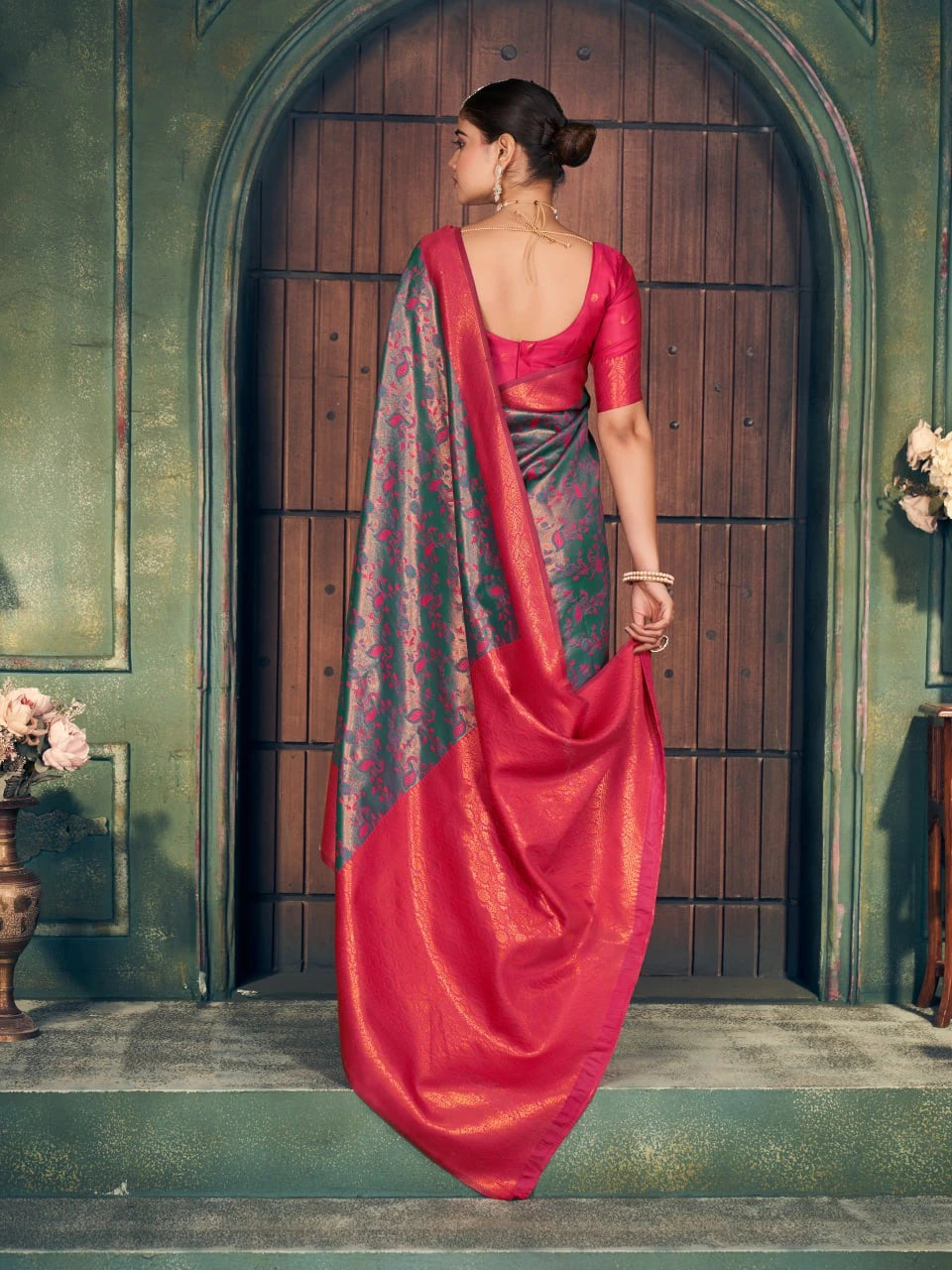 Woman in a red saree with a floral pattern standing in front of a wooden door.