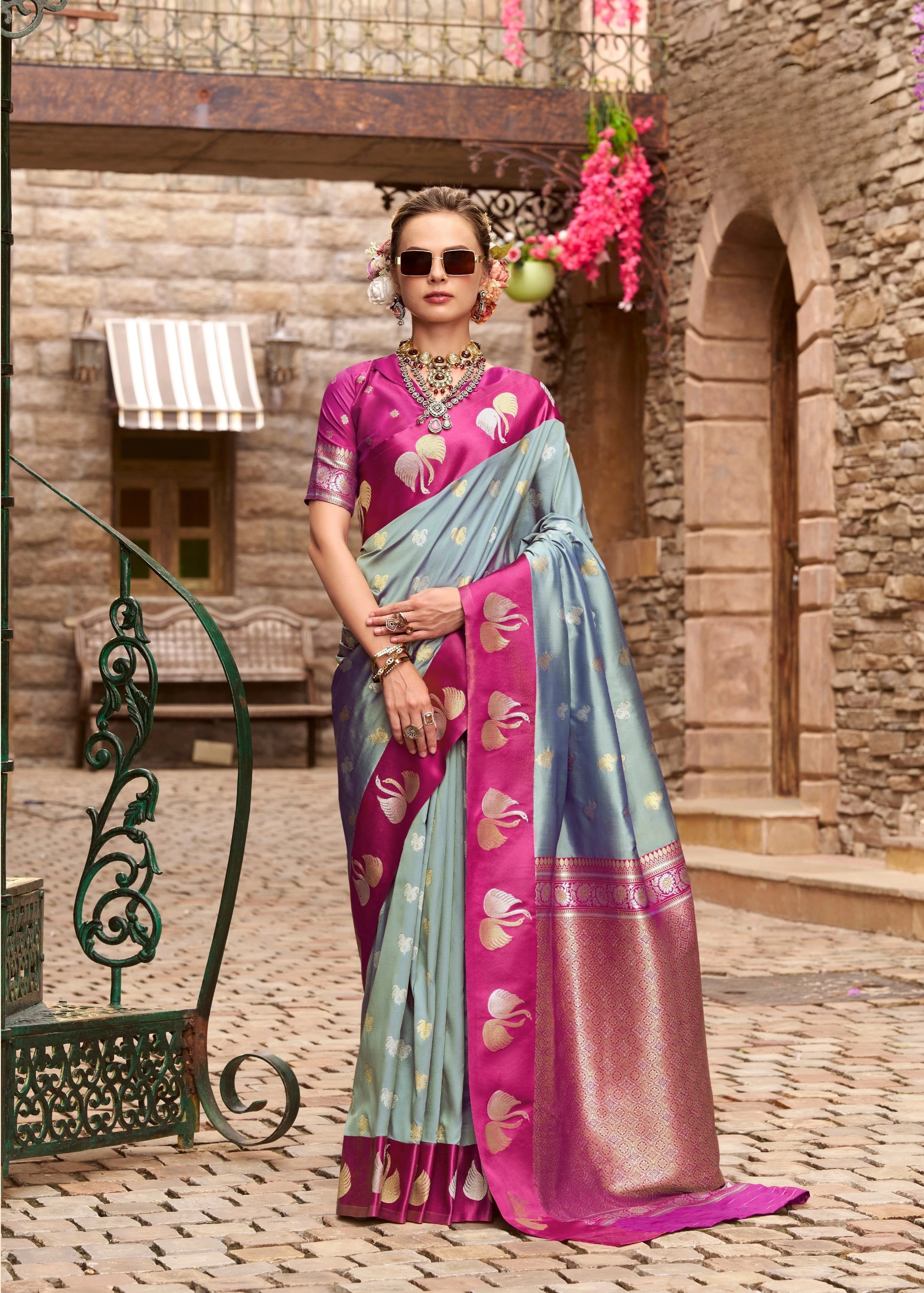 Woman in a pink and blue saree standing in an outdoor setting with stone walls and decorative elements.
