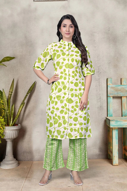 Woman wearing a green floral outfit standing against a textured wall with a plant and wooden chair.