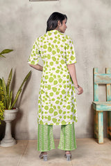 Woman wearing a green and white patterned outfit standing against a neutral wall with a plant and chair in the background.